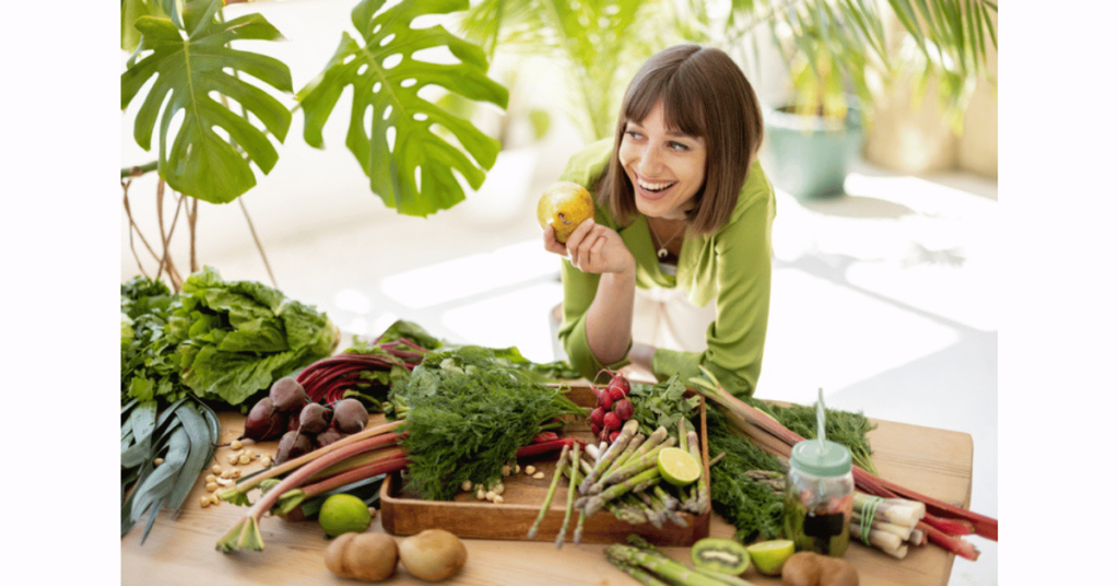 Young cheerful woman by the table full of fresh vegetables, fruits, and greens